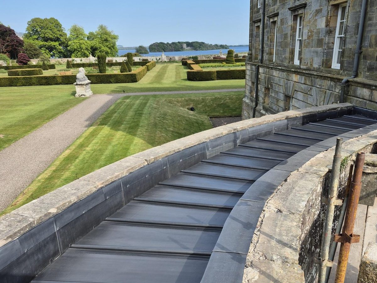 A curved lead lined parapet gutter running along the roof of a stone country house, with formal gardens and a loch beyond