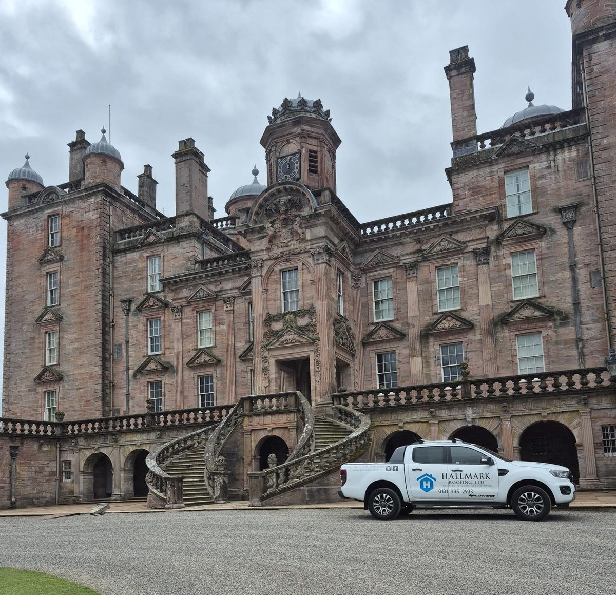 A Hallmark Roofing branded pickup parked in front of a large red sandstone country house with a clock tower and twin stone staircases