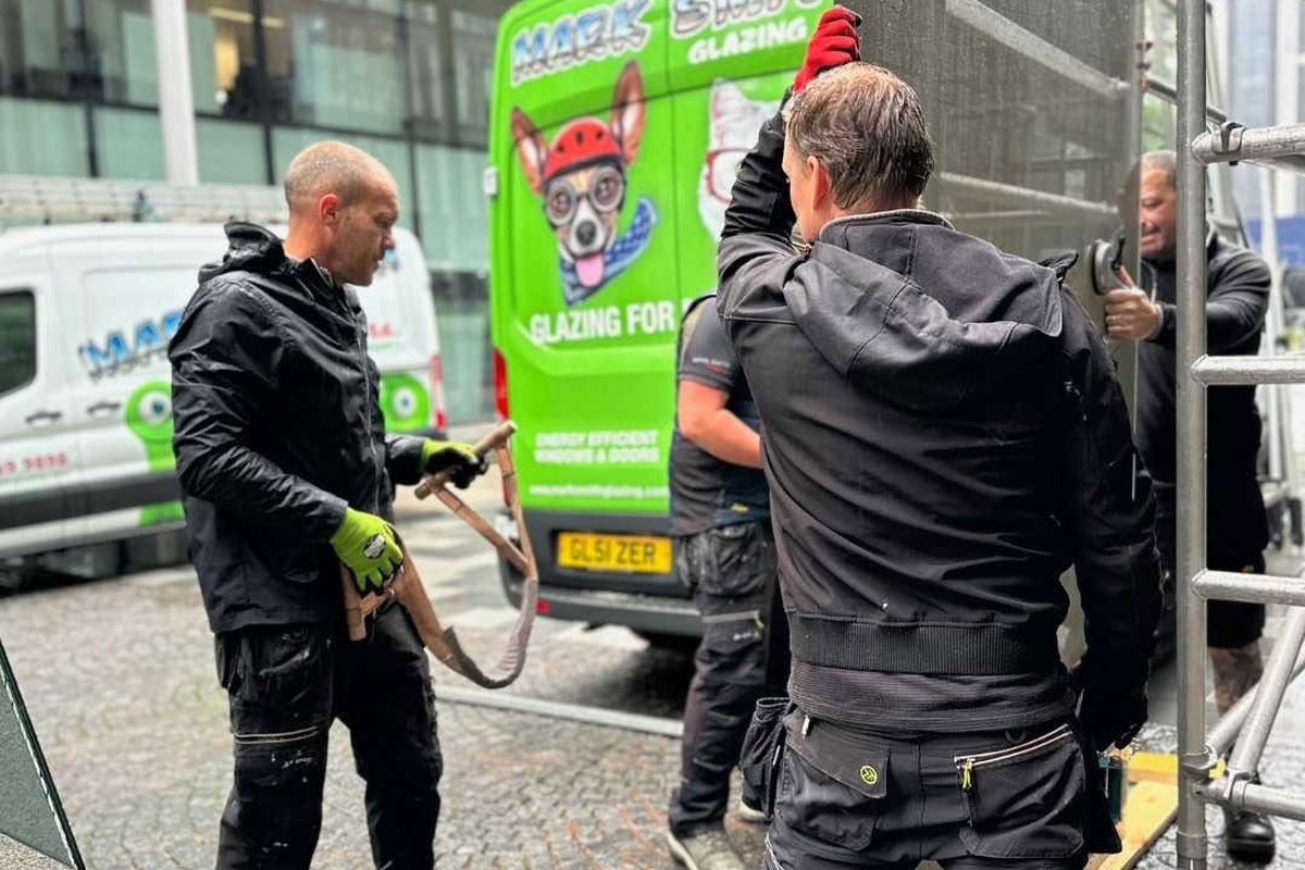 Three Mark Smith Glazing fitters manoeuvring a large glass unit with suction lifters beside a scaffold tower on a wet city centre street, with two company vans behind them